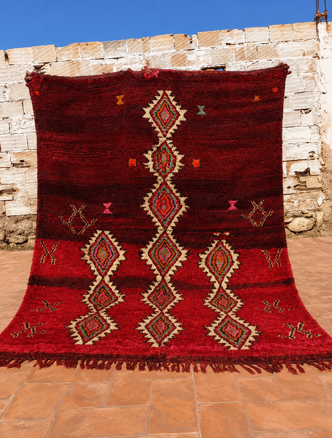 A large red Moroccan wool rug with geometric diamond patterns and fringe, displayed outdoors against a rustic stone wall and terracotta tile floor.