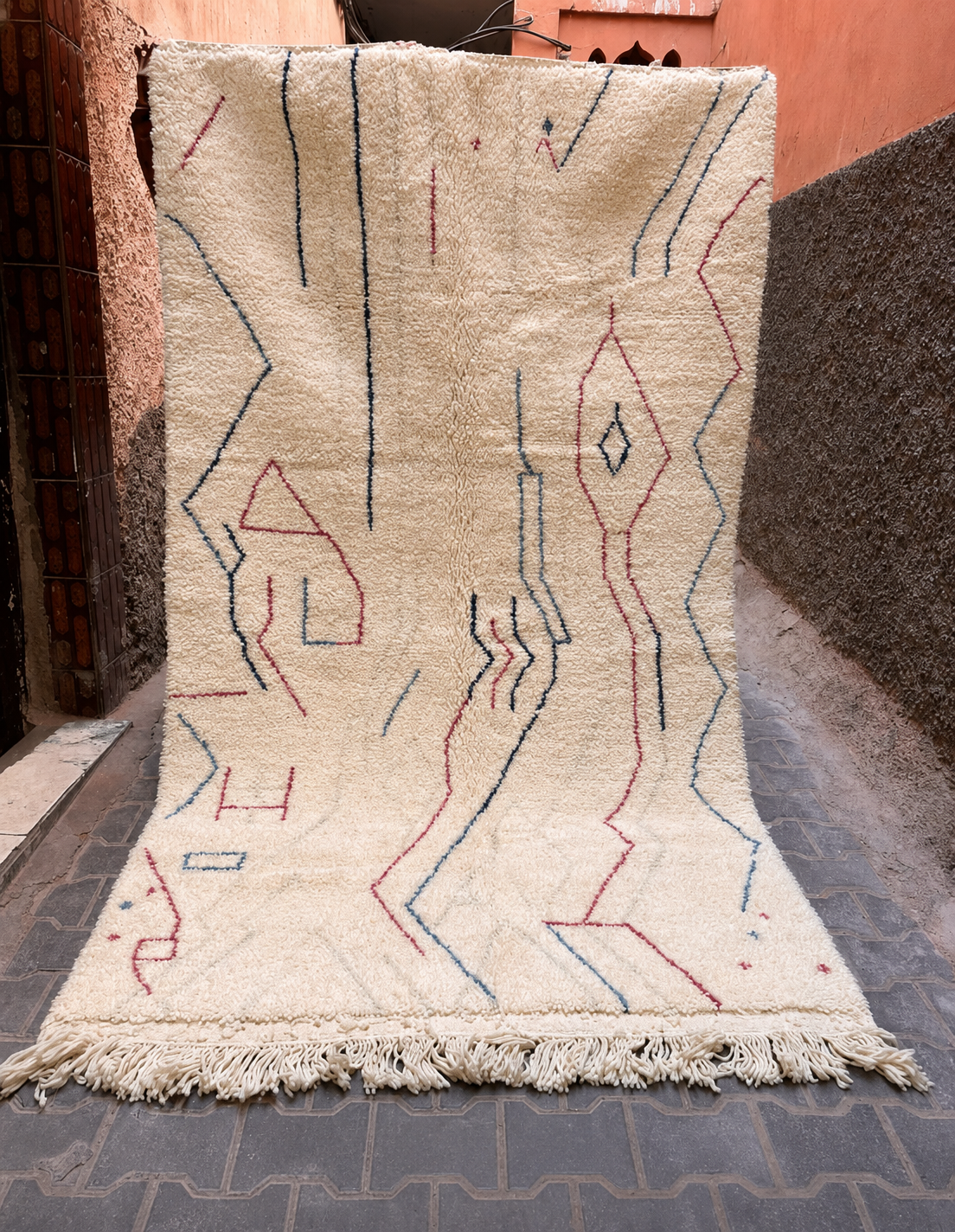 A large off-white Moroccan wool rug with a textured, shaggy surface, featuring hand-drawn geometric patterns in navy blue and muted red. The rug is draped vertically outdoors against a rustic background of a terracotta wall and stone pavement.
